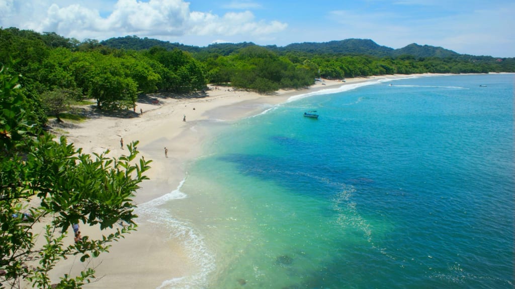 Aerial view of a tropical beach in Costa Rica with white sand, clear blue water, and lush green trees along the shore. A few people and a small boat are visible near the coastline, capturing the serene beauty best seen during the peak wildlife season.